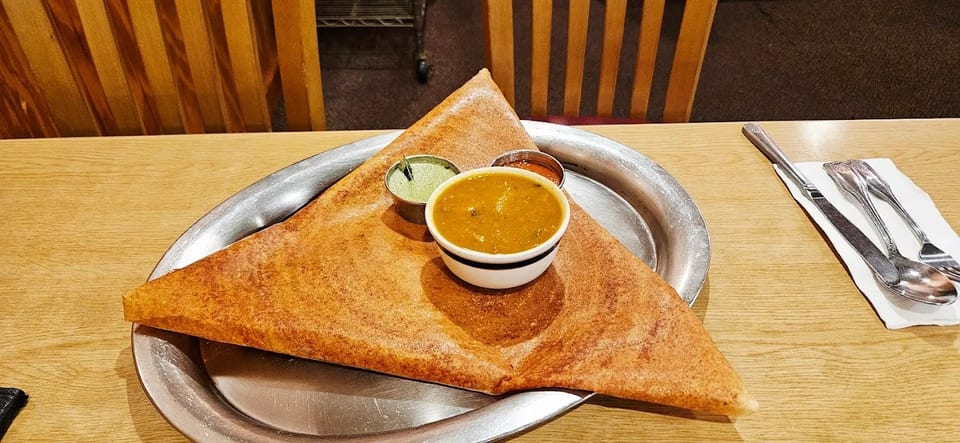A large, triangular dosa on a metal plate with three small bowls of chutney and sambar.