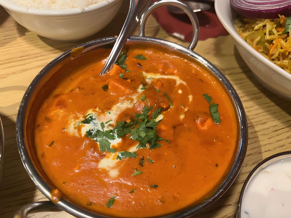 Close-up of a vibrant orange butter chicken curry in a metal bowl, garnished with cream and cilantro.