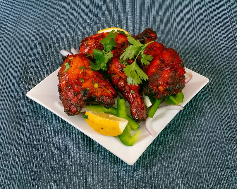 A white plate of vibrant red tandoori chicken, garnished with cilantro, lemon, and bell peppers.