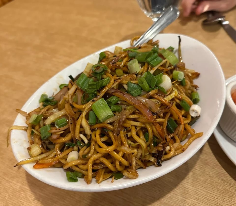 A white oval bowl filled with stir-fried noodles, vegetables, and green onions on a wooden table.