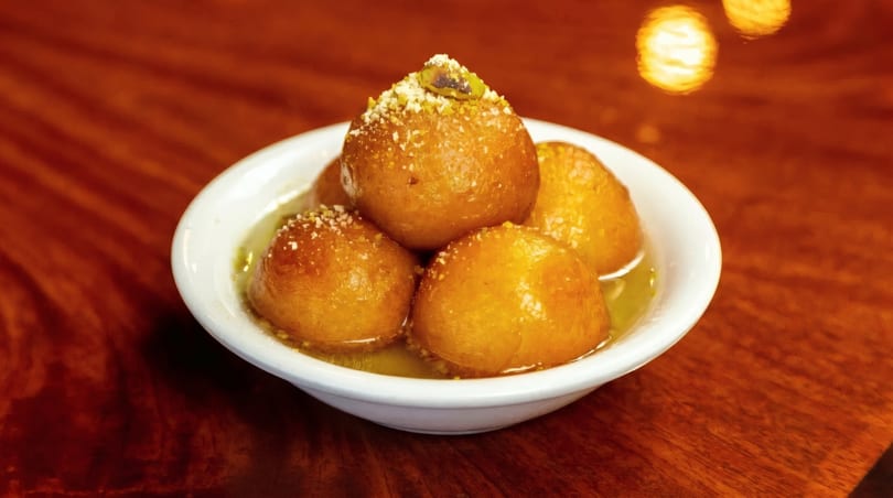 A white bowl filled with golden gulab jamun, soaked in syrup and garnished with nuts.