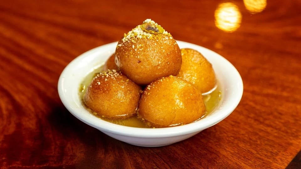 A white bowl filled with golden-brown gulab jamun, garnished with crushed pistachios, on a wooden table.